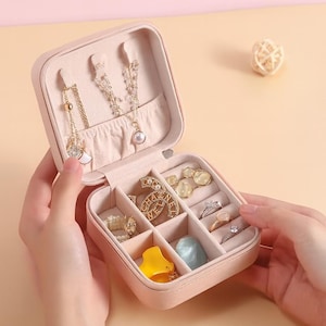 Peut inclure: Une trousse de voyage rose pour bijoux, ouverte pour présenter diverses bagues, colliers et boucles d'oreilles. L'étui est divisé en compartiments pour l'organisation. Les bijoux comprennent des tons or et argent, avec des perles et d'autres éléments décoratifs.