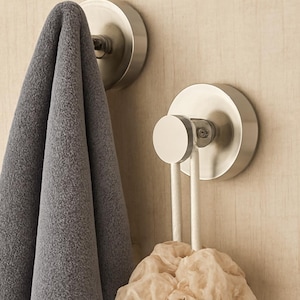 May include: Two brushed nickel wall hooks with a gray towel and a beige bath sponge. The hooks are mounted on a light wood-grain surface. The towel is folded and hanging from one hook.