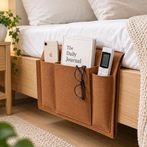 May include: A brown felt bedside storage organizer with three pockets hanging over a wooden bed frame. The pockets contain a smartphone, a book titled The Daily Journal, a pair of black-rimmed eyeglasses, and a white remote control device.