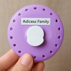Peut inclure: Un objet rond de couleur lavande avec une forme arrondie blanche au centre et le texte "Adicess Family" sur une étiquette blanche. L'objet comporte de petites découpes triangulaires sur le bord.
