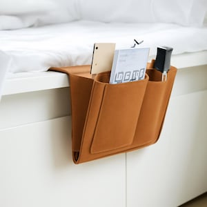 Peut inclure: Un organiseur de chevet en feutre orange brûlé, suspendu au bord d'un cadre de lit blanc. Il comporte trois poches contenant un petit objet en bois, un livre avec le texte "LG 10" et une télécommande noire.