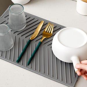 May include: A grey dish drying mat with two clear ribbed glasses, a gold knife and fork with green handles, and a white bowl. The bowl is being held by a person's hand.
