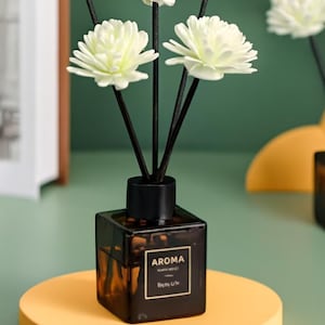 Könnte beinhalten: Aroma-Diffusor mit weißen blütenförmigen Stäbchen und einem gewebten Kugelstäbchen. Die dunkelbraune quadratische Glasflasche steht auf einer gelben kreisförmigen Basis. Die Flasche hat einen schwarzen Deckel und ein Etikett mit der Aufschrift "AROMA".