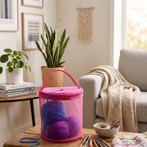 Peut inclure: Un sac de rangement cylindrique en maille rose avec une poignée supérieure, contenant trois pelotes de laine violette. Un fil de laine bleu sort du sac. À proximité se trouvent plusieurs crochets en bois, un bol avec des ciseaux et un textile tricoté.