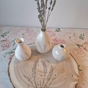 Puede incluir: Tres jarrones de cerámica blanca con ramitas de lavanda, dispuestos sobre una rodaja de madera. Los jarrones tienen un delicado diseño floral en azul. La rodaja de madera se coloca sobre una superficie con estampado floral.