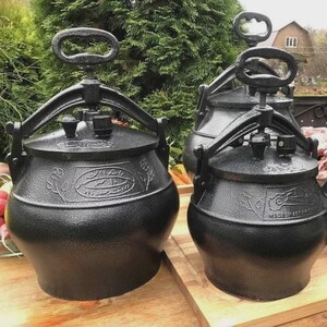 May include: A group of black cast iron cooking pots with handles and pressure release valves are shown on a wooden surface. The pots have a rounded shape with decorative patterns and text. The background features greenery and a building.