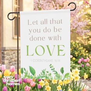Könnte beinhalten: Eine weiße Gartenflagge mit dem Text "Let all that you do be done with LOVE" und "1 Korinther 16,14". Die Flagge hat einen floralen Rand und hängt an einem schwarzen Metallständer. Die Flagge ist von bunten Blumen umgeben.