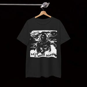 May include: Black t-shirt with a white graphic featuring a hooded figure and the text "DROP A NOISE BOMB". The design has a distressed, vintage aesthetic. The shirt is hanging on a wooden hanger.