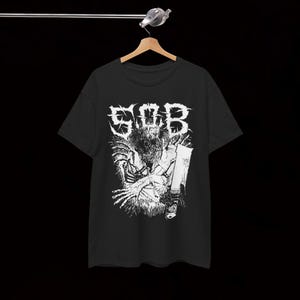 Può includere: T-shirt nero con grafica bianca. La grafica presenta le lettere "SOB" in un font invecchiato sopra un'illustrazione dettagliata di una creatura che tiene una lama. La maglietta è appesa a una gruccia di legno su sfondo nero.