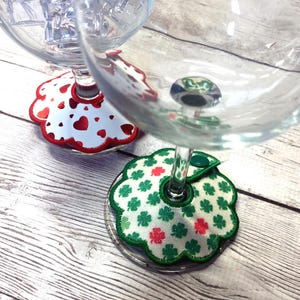 May include: Two wine glasses with decorative coasters. One coaster features a white background with red hearts and a red scalloped edge. The other coaster has a white background with green shamrocks and red accents, with a green scalloped edge.