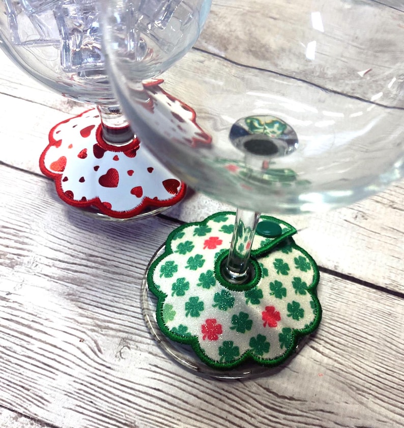 May include: Two wine glasses with decorative coasters. One coaster features a white background with red hearts and a red scalloped edge. The other coaster has a white background with green shamrocks and red accents, with a green scalloped edge.