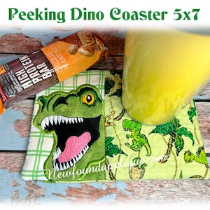 May include: A square coaster with a green dinosaur design, a yellow mug, and a protein bar. The coaster features a winking dinosaur with a pink tongue and the text "Peeking Dino Coaster 5x7". The protein bar is chocolate peanut butter flavored.