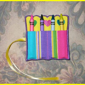 May include: A fabric crayon holder with four crayons. The holder features a striped pattern in turquoise, purple, yellow, and pink. The top edge is yellow with heart and green accents. A yellow ribbon is attached.