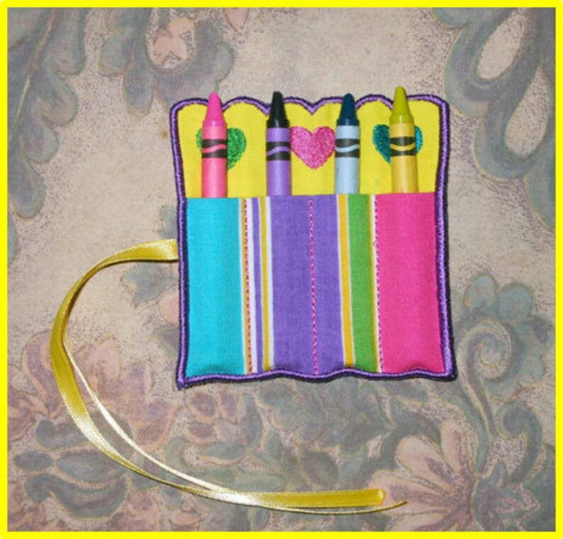 May include: A fabric crayon holder with four crayons. The holder features a striped pattern in turquoise, purple, yellow, and pink. The top edge is yellow with heart and green accents. A yellow ribbon is attached.