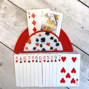 May include: A red playing card holder with a white patterned center, holding two playing cards. The holder is shaped like a half-circle. A deck of playing cards is below the holder.
