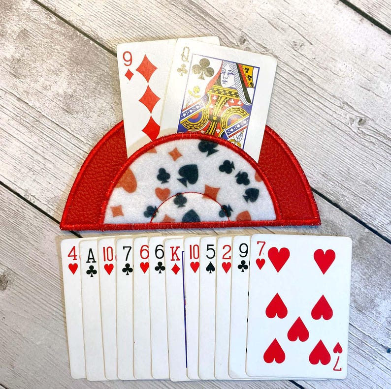 May include: A red playing card holder with a white patterned center, holding two playing cards. The holder is shaped like a half-circle. A deck of playing cards is below the holder.