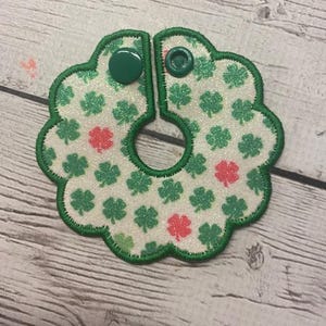 May include: A two-piece, clover-themed bib with green trim and two green snap closures. The bib features a white background with green and red glitter shamrocks.
