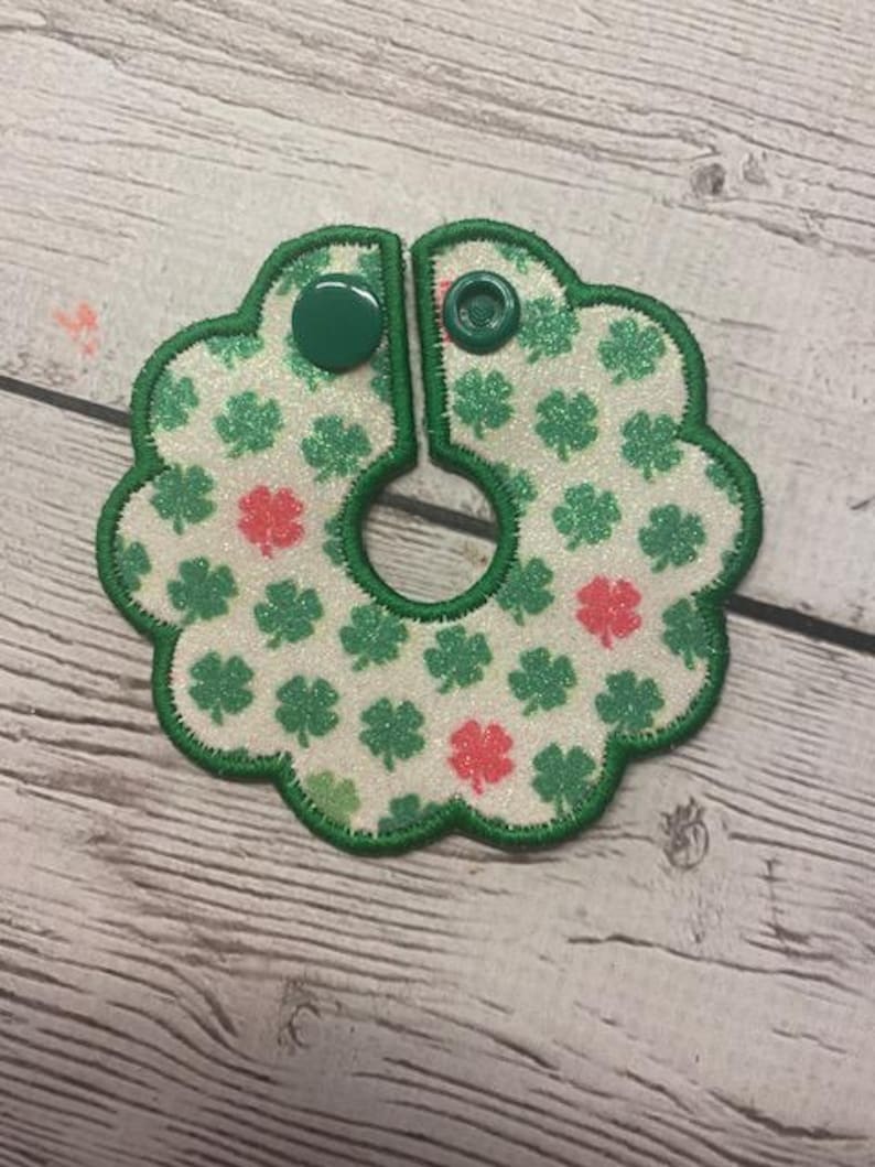 May include: A two-piece, clover-themed bib with green trim and two green snap closures. The bib features a white background with green and red glitter shamrocks.