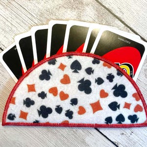 May include: A white felt card holder with a red border, decorated with orange and black card suit symbols. The holder is filled with black and red playing cards. The background is a light wood grain.
