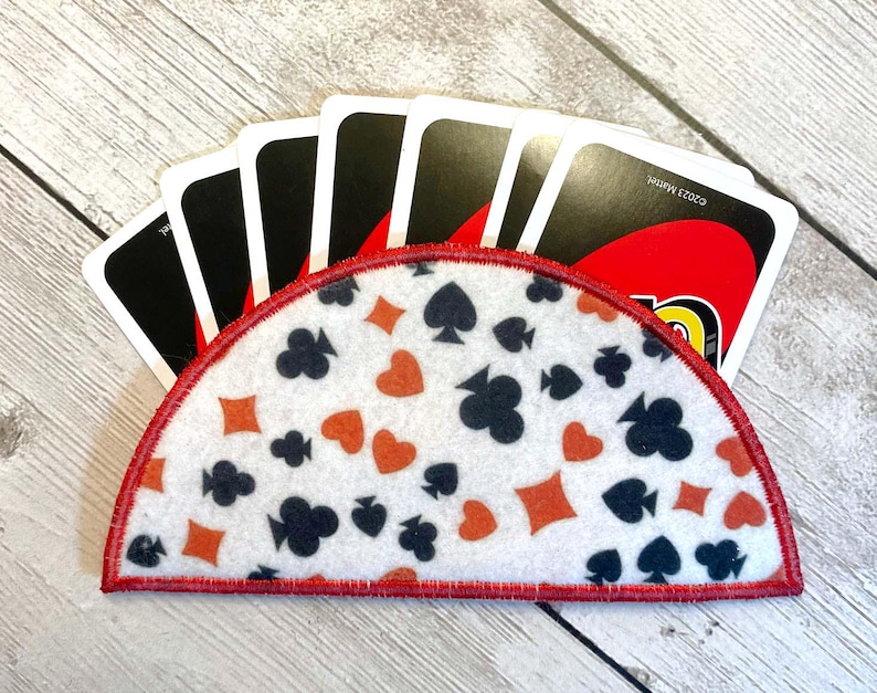 May include: A white felt card holder with a red border, decorated with orange and black card suit symbols. The holder is filled with black and red playing cards. The background is a light wood grain.