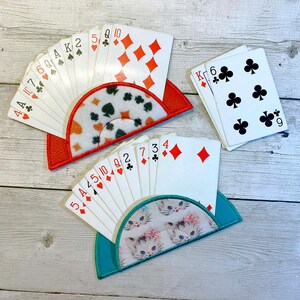 May include: Three playing card holders with decks of cards. One holder is red with a patterned insert, another is teal with a kitten print, and the third is a stack of cards. The cards show suits of diamonds, clubs, spades, and hearts.