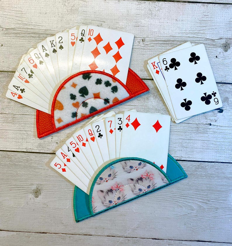 May include: Three playing card holders with decks of cards. One holder is red with a patterned insert, another is teal with a kitten print, and the third is a stack of cards. The cards show suits of diamonds, clubs, spades, and hearts.