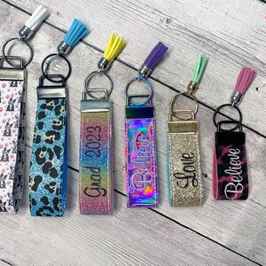 May include: Seven colorful keychains with different designs and tassels. The keychains feature various patterns, including a black and white dog print, a blue and black leopard print, a rainbow glitter design, a holographic design with the word "Believe", a gold glitter design with the word "Love", and a black and pink tie-dye design with the word "Believe". One keychain has the text "Grad 2023" on it.