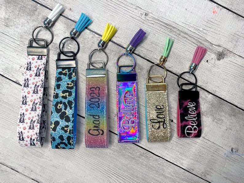 May include: Seven colorful keychains with different designs and tassels. The keychains feature various patterns, including a black and white dog print, a blue and black leopard print, a rainbow glitter design, a holographic design with the word "Believe", a gold glitter design with the word "Love", and a black and pink tie-dye design with the word "Believe". One keychain has the text "Grad 2023" on it.