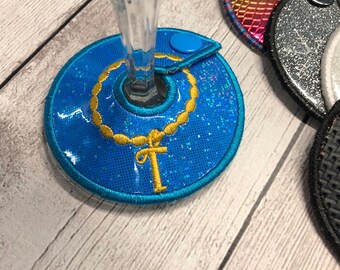 May include: A blue glittery drink coaster with a gold embroidered ankh and chain design. The coaster has a small blue button and is designed to hold a glass. Other coasters are visible in the background.