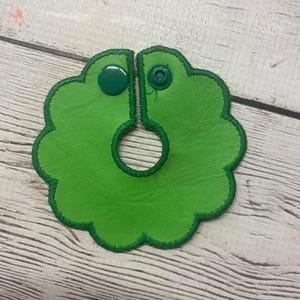 May include: A bright green leather bib with a scalloped edge and a dark green stitched border. The bib has a snap closure and a circular opening in the center. The bib is designed to be worn around the neck.