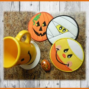 May include: Four Halloween-themed coasters with embroidered designs. The coasters feature a pumpkin, a mummy, a candy corn, and a smiling face. The coasters are on a brown surface with a yellow mug and a small candy.