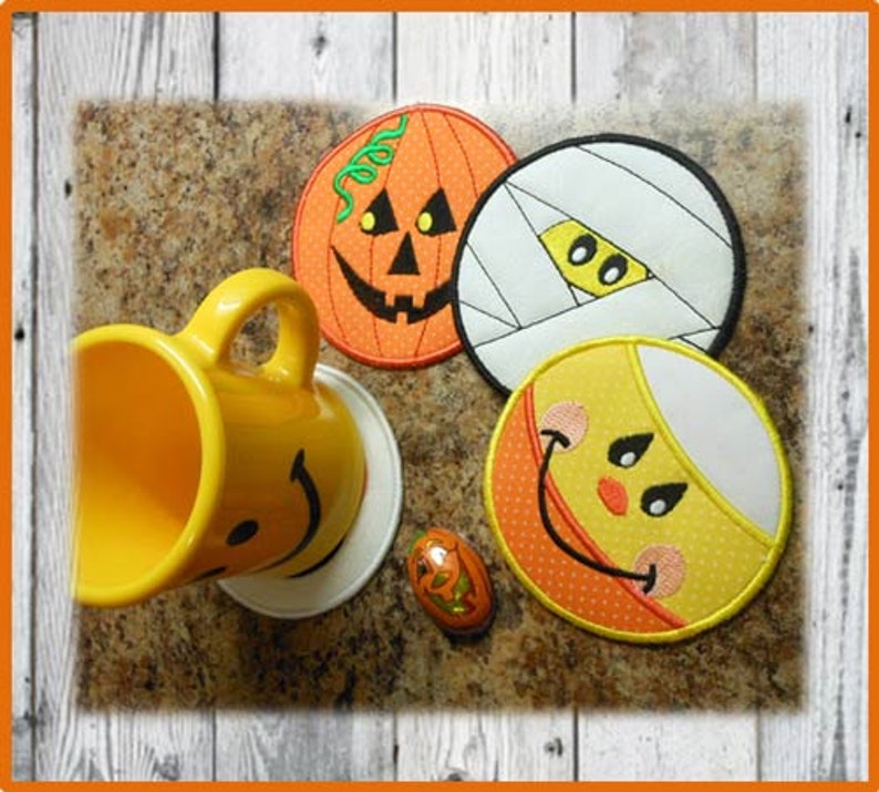 May include: Four Halloween-themed coasters with embroidered designs. The coasters feature a pumpkin, a mummy, a candy corn, and a smiling face. The coasters are on a brown surface with a yellow mug and a small candy.