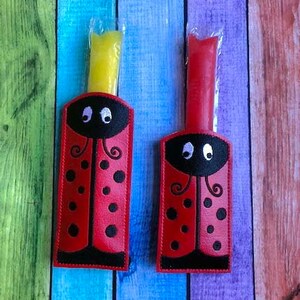 May include: Two red ladybug-shaped holders with black spots and a black head, each holding a frozen treat. One treat is yellow, the other is red. The holders are on a colorful wooden background.