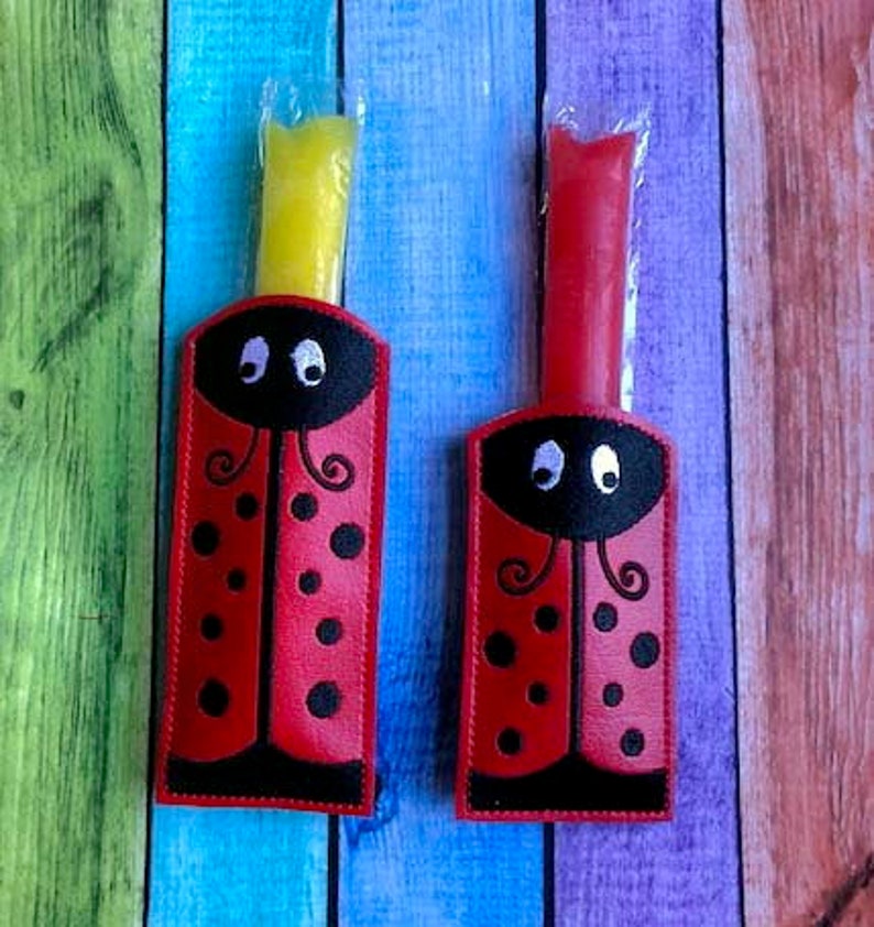 May include: Two red ladybug-shaped holders with black spots and a black head, each holding a frozen treat. One treat is yellow, the other is red. The holders are on a colorful wooden background.