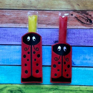 May include: Two ladybug-themed popsicle holders, one with yellow and the other with red popsicles. The holders are red with black accents, black spots, and embroidered eyes. The background is a colorful wooden surface.