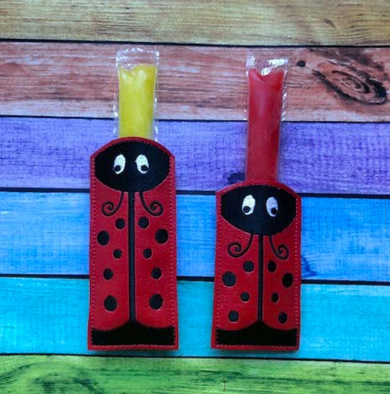 May include: Two ladybug-themed popsicle holders, one with yellow and the other with red popsicles. The holders are red with black accents, black spots, and embroidered eyes. The background is a colorful wooden surface.