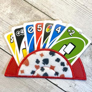 May include: A red card holder with a white and red patterned design, holding a hand of Uno cards. The cards are yellow, blue, red, and green, with various numbers and symbols. The holder is made of a textured material.