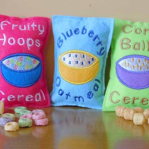 May include: Three felt pouches with embroidered cereal box designs. The pouches are pink, blue, and green. The pink pouch says "Fruity Hoops Cereal", the blue pouch says "Blueberry Oatmeal", and the green pouch says "Corn Balls Cereal". There are cereal pieces scattered in front of the pouches.