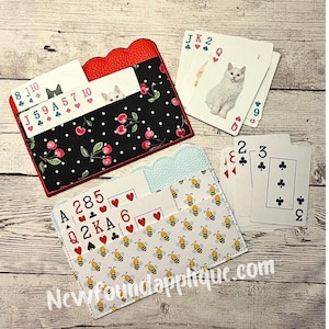 Peut inclure: Deux étuis pour cartes à jouer avec différents motifs. L'un est noir avec des cerises rouges, l'autre est blanc avec des abeilles jaunes. Plusieurs cartes à jouer sont visibles, avec le texte "Newfoundapplique.com".