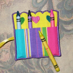 May include: A vibrant crayon holder with a yellow, scalloped top, embroidered hearts, and striped pockets. It holds four crayons in shades of blue, purple, and pink. A yellow crayon is placed diagonally across the front.