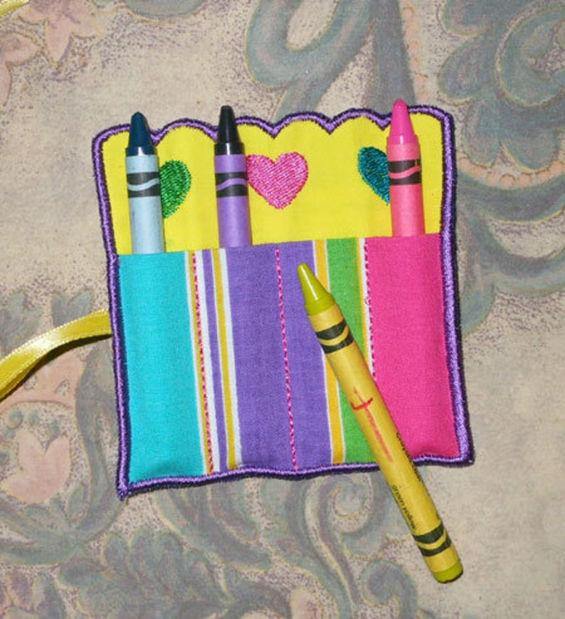 May include: A vibrant crayon holder with a yellow, scalloped top, embroidered hearts, and striped pockets. It holds four crayons in shades of blue, purple, and pink. A yellow crayon is placed diagonally across the front.