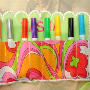 May include: A fabric marker holder with a colorful floral pattern and a scalloped edge. The holder contains seven markers in various colors, with the name "ASHLEY" embroidered in green. A green ribbon is attached for closure.