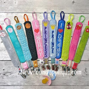 May include: A selection of brightly coloured pacifier clips. Designs include sports balls, hearts, and text such as "Mommy" and "Princess." Each clip has a metal clasp and a loop for attaching to a pacifier or toy.
