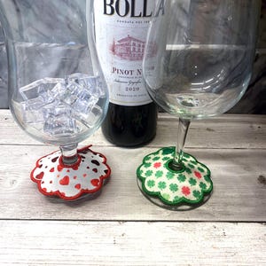 May include: A bottle of Bolla Pinot Noir wine is flanked by two wine glasses, one filled with ice cubes. Decorative coasters with heart and shamrock designs sit beneath the glasses. The image is set on a light wood surface.