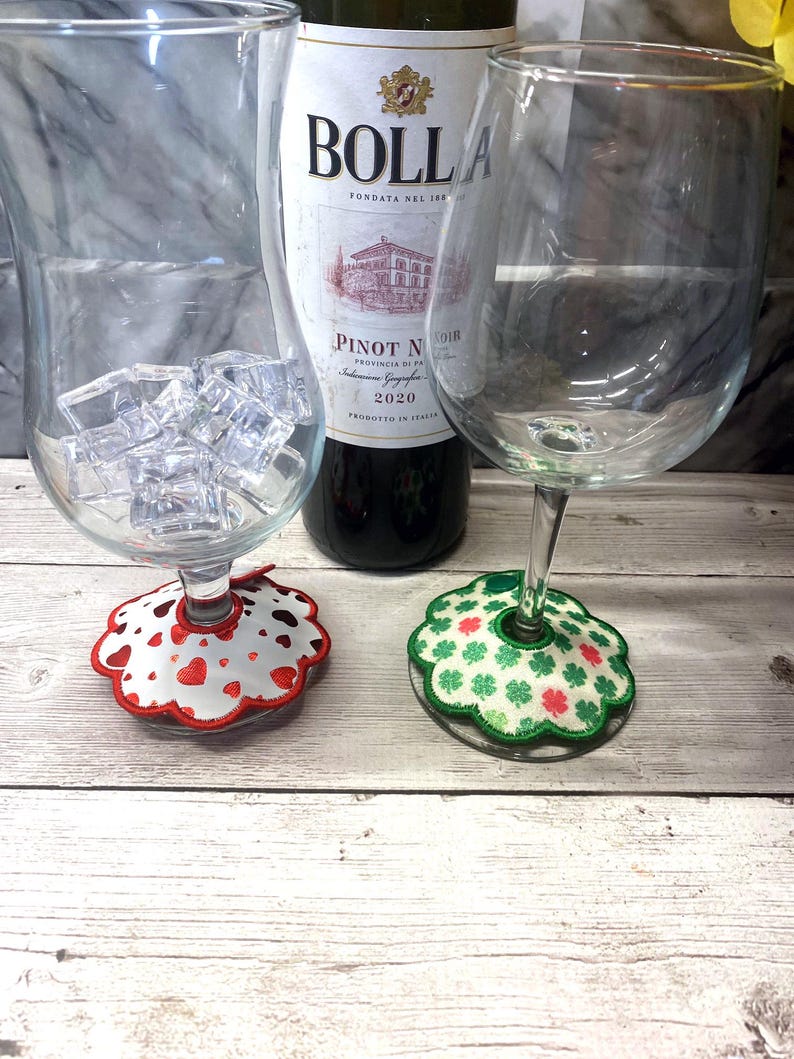 May include: A bottle of Bolla Pinot Noir wine is flanked by two wine glasses, one filled with ice cubes. Decorative coasters with heart and shamrock designs sit beneath the glasses. The image is set on a light wood surface.