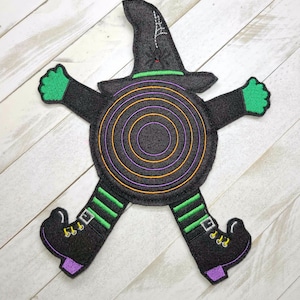 May include: A felt witch applique with a black hat, green arms, and purple boots. The witch's body is a black circle with orange and purple concentric circles.
