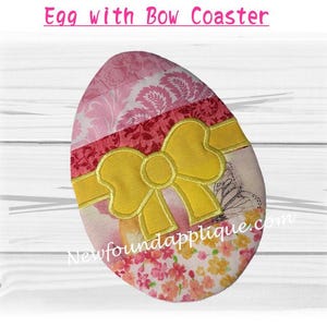 Puede incluir: Un posavasos en forma de huevo con un diseño de lazo amarillo. El posavasos presenta un mosaico de patrones rosas, rojos y florales. El texto "Egg with Bow Coaster" está en la parte superior. El sitio web "Newfoundapplique.com" está en la parte inferior.