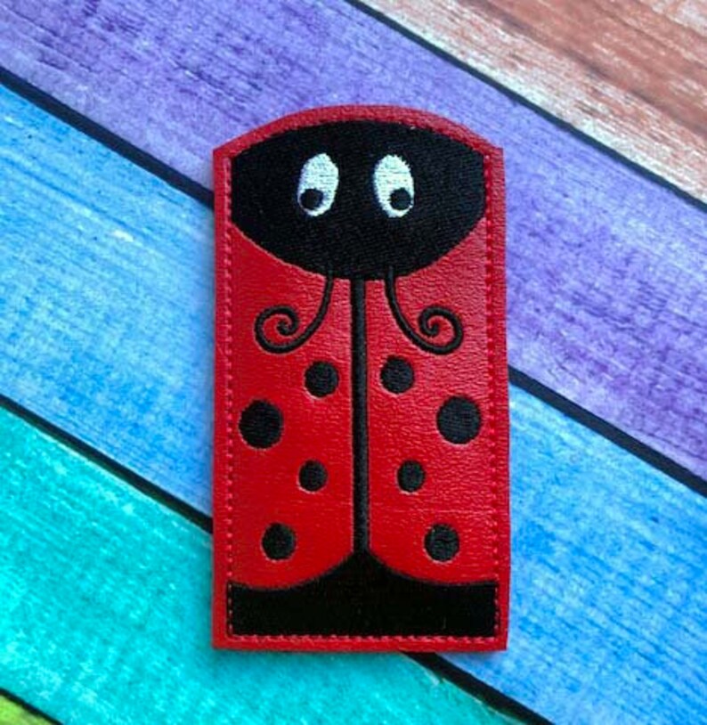 May include: A red ladybug-shaped applique with black spots and a black head. The applique has white eyes and is made of a smooth material. The ladybug design is a whimsical and playful element.
