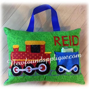 May include: A green pillow with a red and yellow train embroidered on it. The pillow has a blue handle and a pocket with the name "REID" and the website "newfoundapplique.com" embroidered on it.