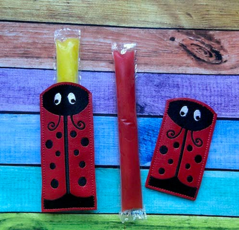 May include: Red ladybug-themed popsicle holders with black accents and a yellow popsicle. A red popsicle is next to the holder. The ladybug design includes black spots and a black head with white eyes.